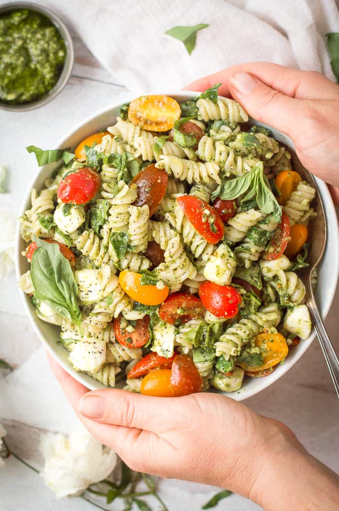 Pesto veggie pasta salad in a white bowl with a spoon. Hands are holding the bowl. There is a white napkin and cup of pesto next to it.