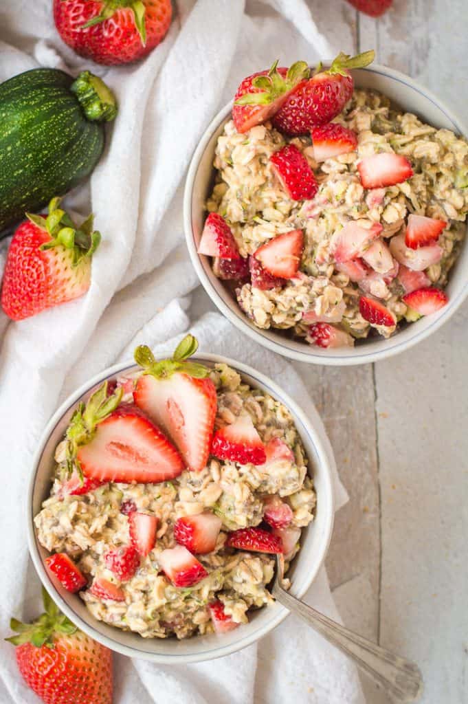 Two bowls of zucchini strawberry overnight oats on a table with a white napkin and berries.