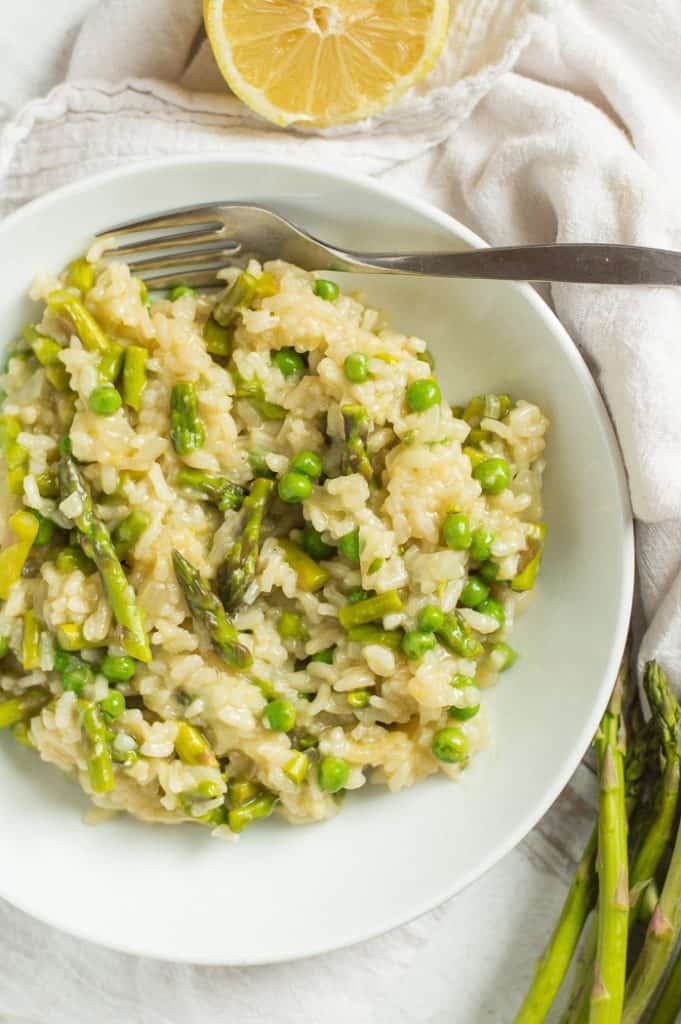 A white bowl of asparagus and pea risotto on a table with a fork. There is fresh asparagus and a lemon on the table with it.