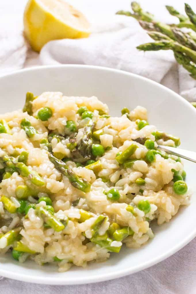 A bowl of asparagus and pea risotto on a table with a white napkin. There is a fork on the bowl and blurred lemon and asparagus in the background. 