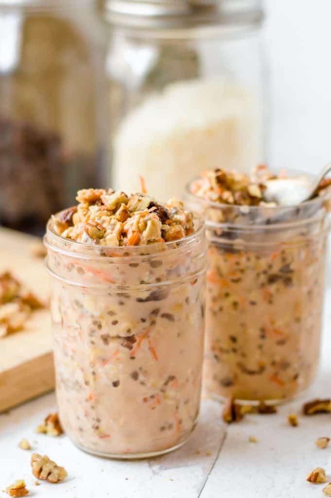 Carrot cake overnight oats in two jelly jars. There are nuts scattered around on the table a blurred jars in the background.