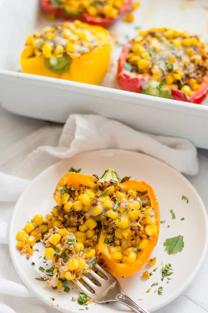 A corn quinoa stuffed pepper on a plate with a fork taking a bite out of it. The tray of stuffed peppers is in the background and there is a napkin under the plate.