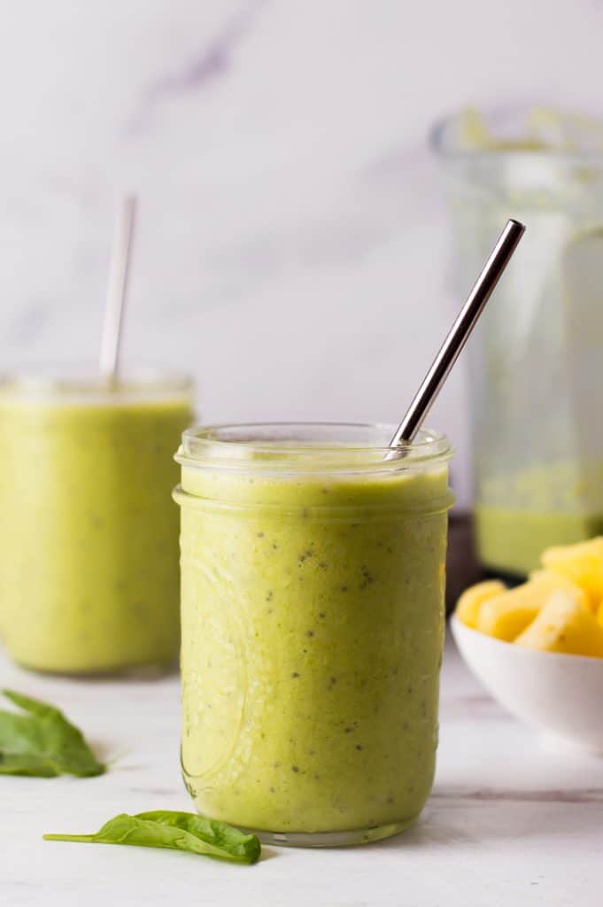 two glass jars filled with green smoothie and metal straws, blender and pineapple in the background

