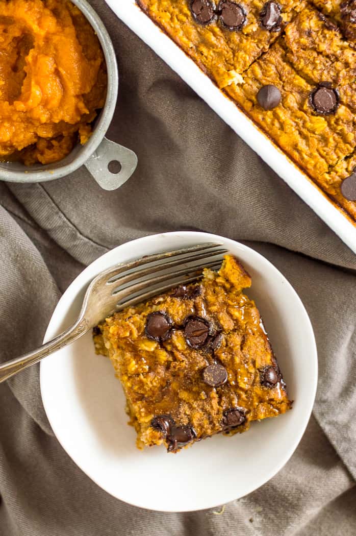 A slice of pumpkin baked oatmeal in a bowl with a fork. There is the baking tray of oatmeal and a measuring cup of pumpkin next to it.