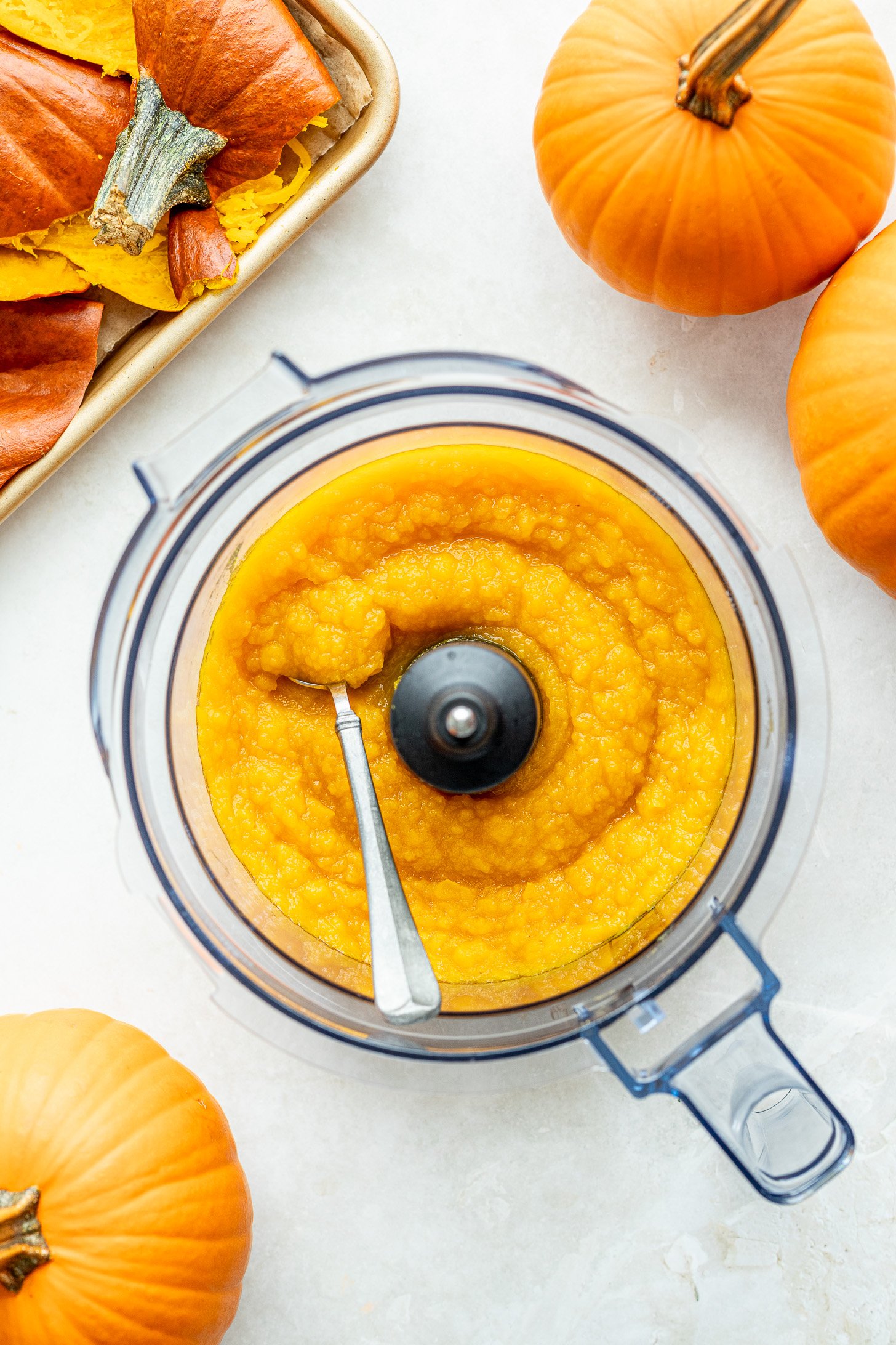 Pumpkin puree in a food processor with a spoon and pumpkins around it on a table.