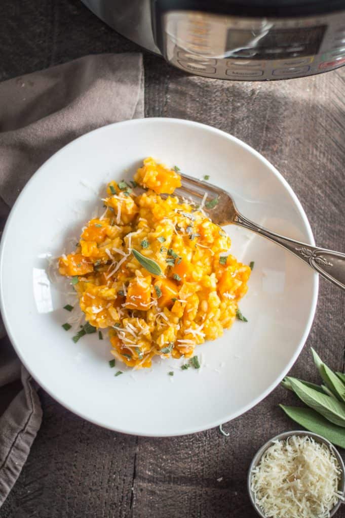 Instant Pot butternut squash risotto in a bowl on a table. The Instant Pot is next to the bowl and their is a measuring cup of shredded cheese and sage next to it.