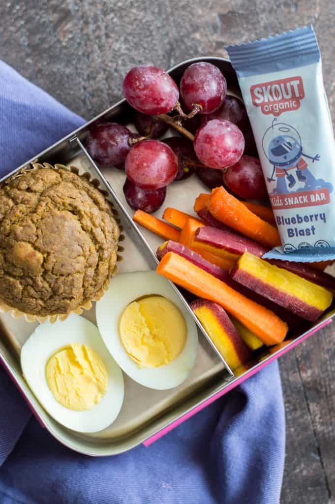 A metal lunch box with two compartments. One compartment has with a muffin and hard boiled eggs. The other compartment has grapes, carrot sticks and a Skout Organic bar.
