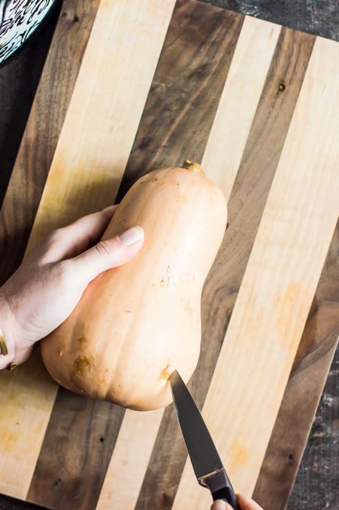 A butternut squash on a cutting board. A hand is holding it and is puncturing the squash with a knife.
