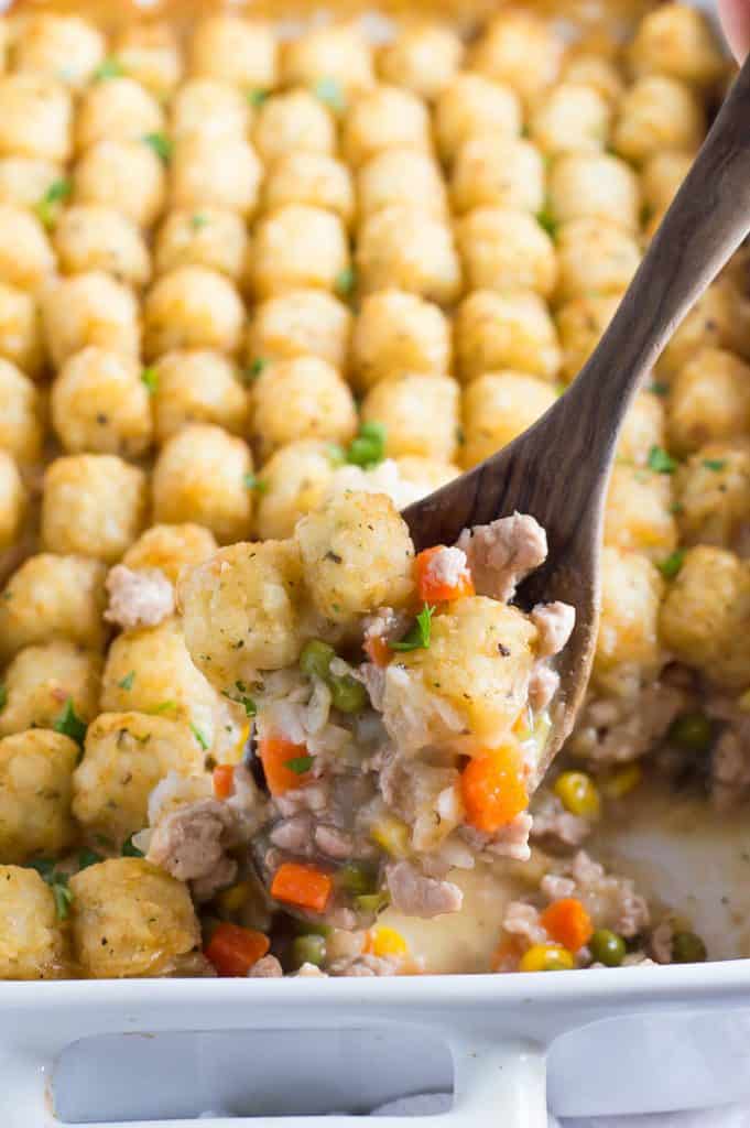 A wooden spoon taking a scoop of veggie-loaded tater tot casserole out of the dish.