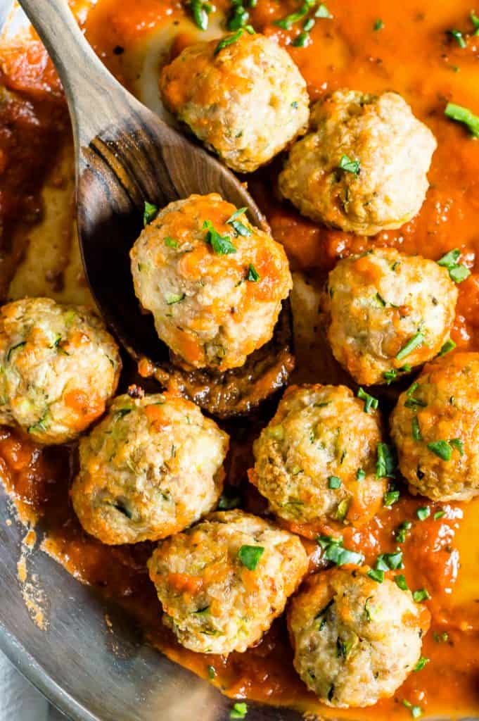 Turkey zucchini meatballs in a pan with sauce and a wooden spoon is scooping one up.