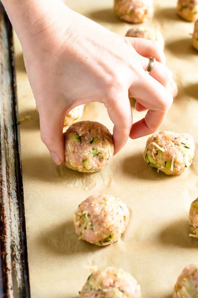 Turkey zucchini meatballs on a baking sheet. A hand is placing one meatball into place.