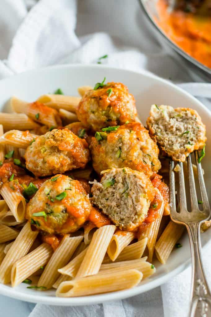 Turkey zucchini meatballs in a bowl with pasta and sauce. A fork has cut a meatball in half and has half on the prongs.