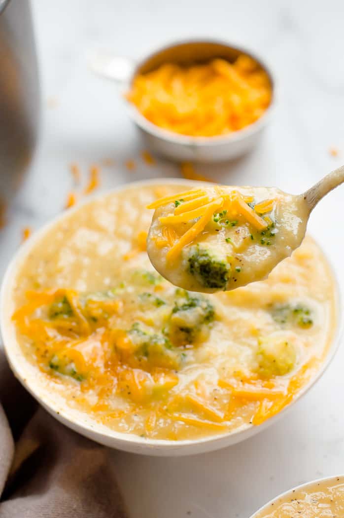 Healthy broccoli cheddar soup in a bowl. A spoon is taking a scoop of soup and there is a cup of shredded cheese blurred in the background.