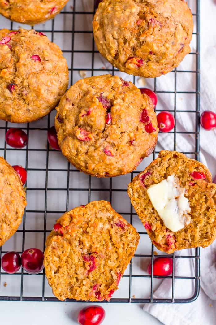 Healthy cranberry orange carrot muffins on a cooling rack with fresh cranberries. One muffin is cut open and has butter spread on one half of it.