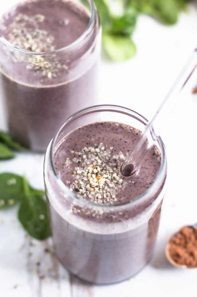 Spinach Cacao smoothie in a glass topped with some hemp hearts. It has a glass straw in it and there is another smoothie and some spinach blurred in the background.