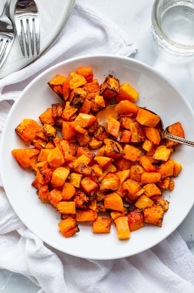 Air fryer butternut squash in a white bowl with a serving spoon. There is a plate with forks and a glass of water next to the bowl of squash.