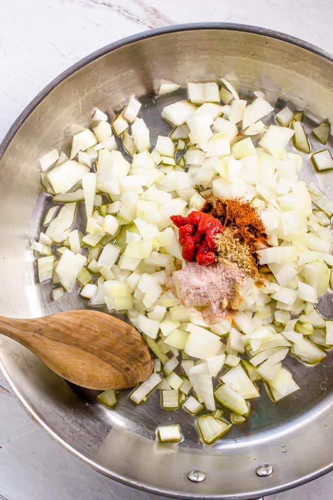 Onions, tomato paste, and seasoning in a stainless steel skillet with a wooden spoon.
