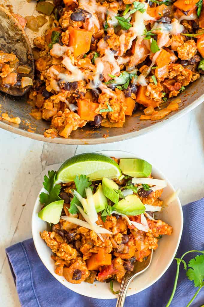 A serving of sweet potato turkey taco skillet in a bowl with avocado, limes, cheese and cilantro on a table with a blue napkin. The skillet is next to the bowl on the table.