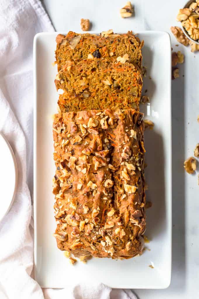 Carrot cake bread on a white plate with two slices cut from it. There is a white napkin and walnuts scattered around the plate.