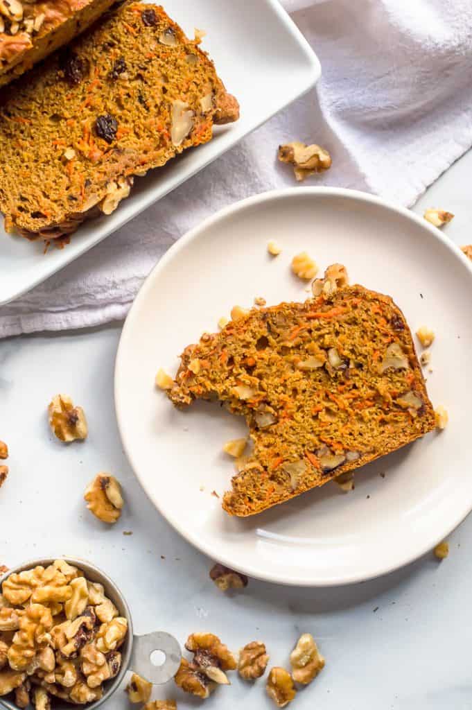 A slice of carrot cake bread on a plate with a bite taken out of it. There is another slice on a plate next to it as well as a measuring cup of walnuts.