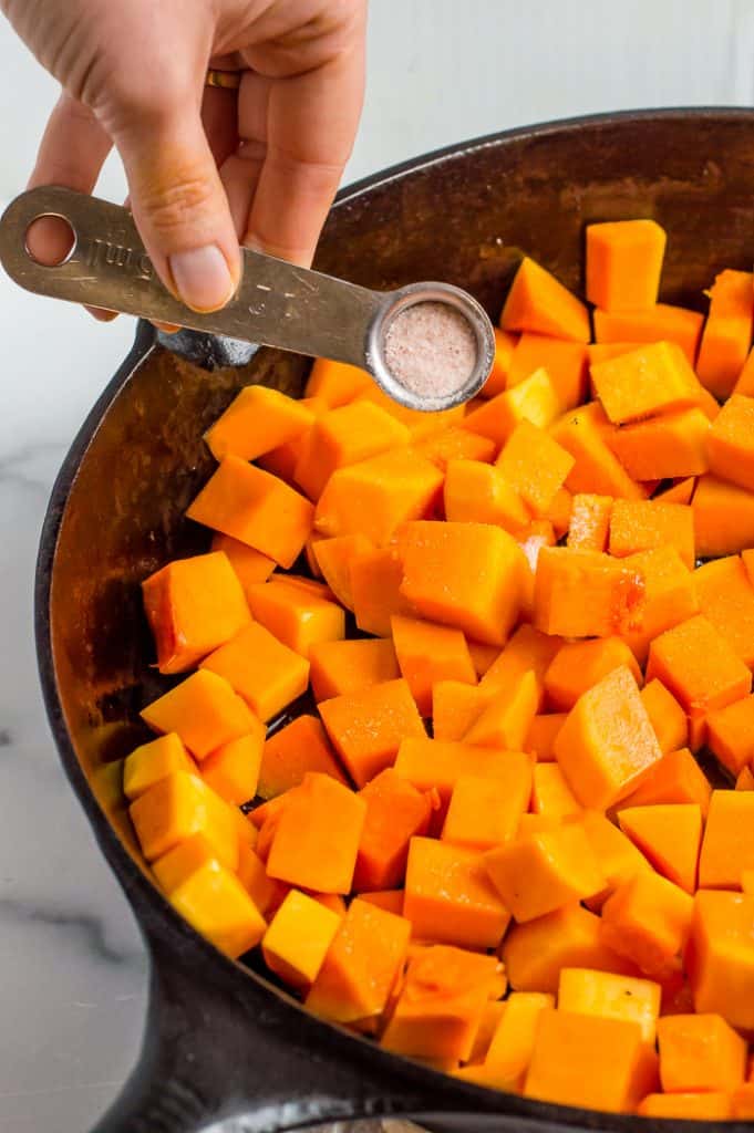 Butternut squash in a cast iron skillet. A measuring spoon is pouring salt over it. 