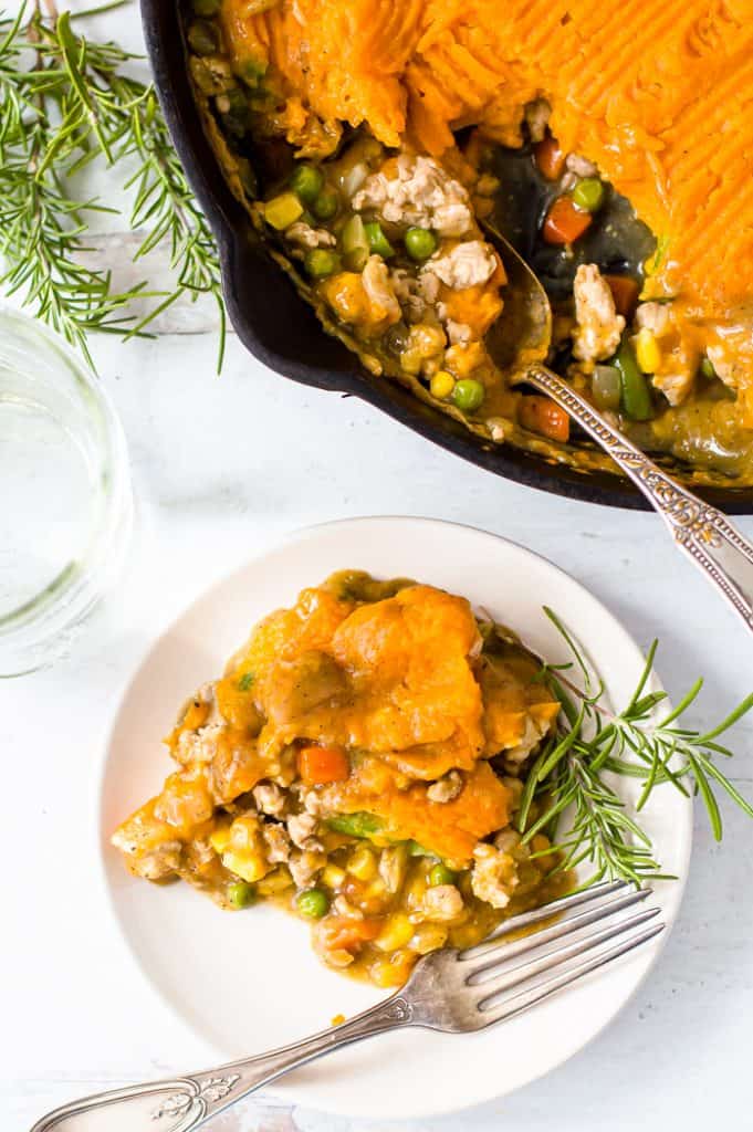 A serving of turkey sweet potato shepherd's pie  on a plate with fresh rosemary and a fork. The skillet with the rest of the pie is on the table next to it. 
