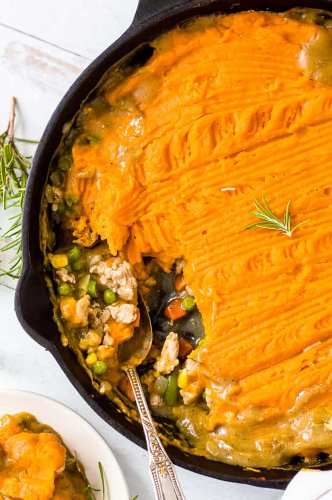 Turkey sweet potato Shepherd's pie in a cast iron  with a spoon in it. A serving has been scooped out and there is a piece of fresh rosemary on top of the rest of the pie. 