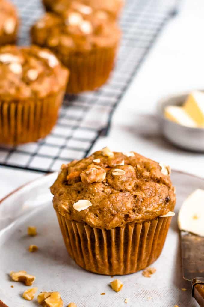 A carrot banana muffins on a plate with nuts scattered around and a butter knife with butter on it. The cooling rack with muffins and a small tin of butter is blurred in the background