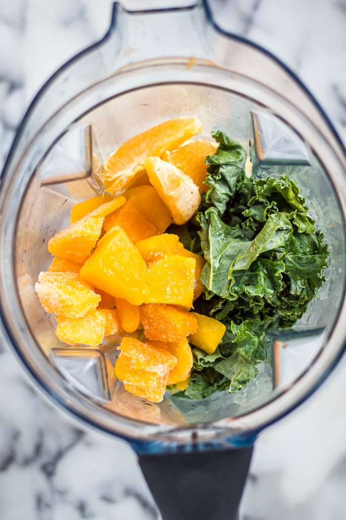A blender with kale, mango and and orange in it before being blended into a smoothie.