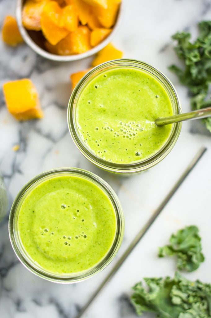 Two glasses of mango kale smoothie on a table with fresh kale and a bowl of mango next to them. One glass has a metal straw in it and there is another straw on the table.