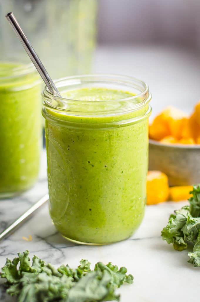 A mason jar filled with mango kale smoothie. There is a straw in the glass and there are leaves of kale and a blurred bowl of frozen mango in the background. 