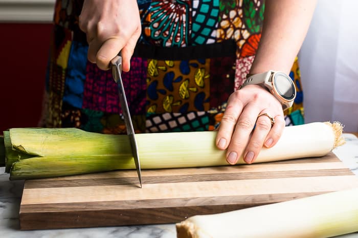 Person holding onto a whole leek on a wooden cutting board with one hand and slicing the green leaves off the top of the leek with a chef's knife in the other hand.