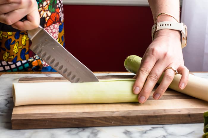 Person holding onto a leek on a wooden cutting board with one hand and slicing the leek in half lengthwise with a chef's knife in the other hand.