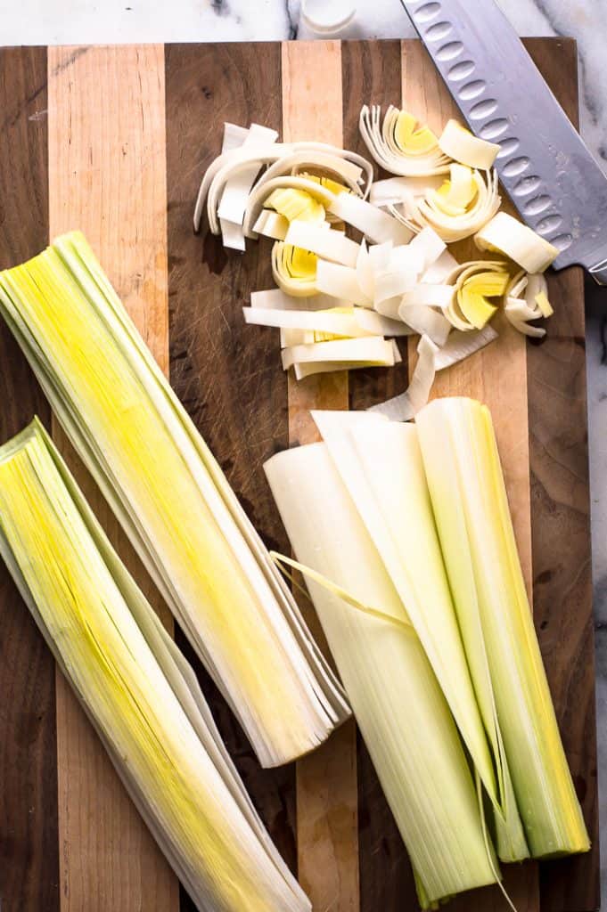 Leeks on a cutting board with a chef's knife in the upper right corner. The leeks are cut in half lengthwise as well as cut into half moon shapes.