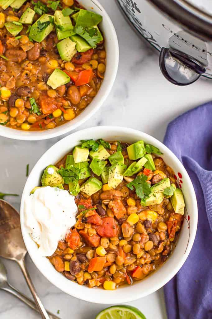 Slow cooker veggie chili in two white bowls, topped with diced avocado, sour cream and fresh cilantro. There are two spoons and a blue napkin on the table next to the bowls