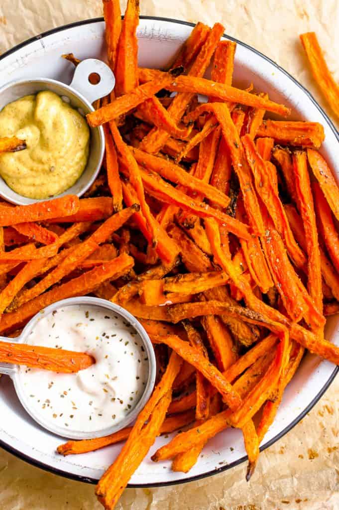 A plate of carrot fries with a small dish of ranch dressing and mustard with a carrot fry in it.