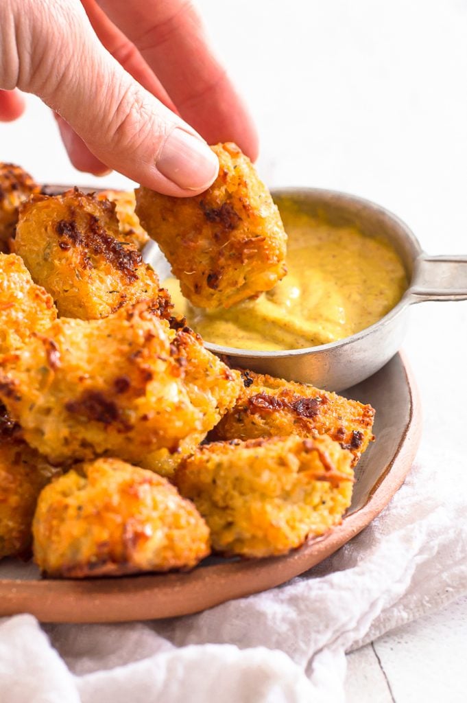 A hand dipping a cauliflower tot in a cup of mustard 