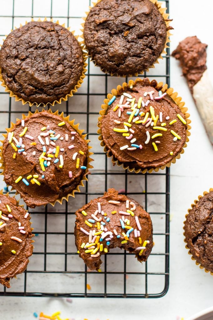 chocolate sweet potato cupcakes on a cooling rack. Some have frosting and sprinkles, others do not. One cupcake has a bite taken out of it.