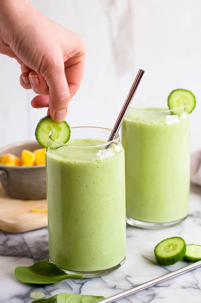 A hand putting a cucumber slice on the edge of a glass with pineapple cucumber smoothie. The glass has a metal straw and there is another cup of smoothie behind it.