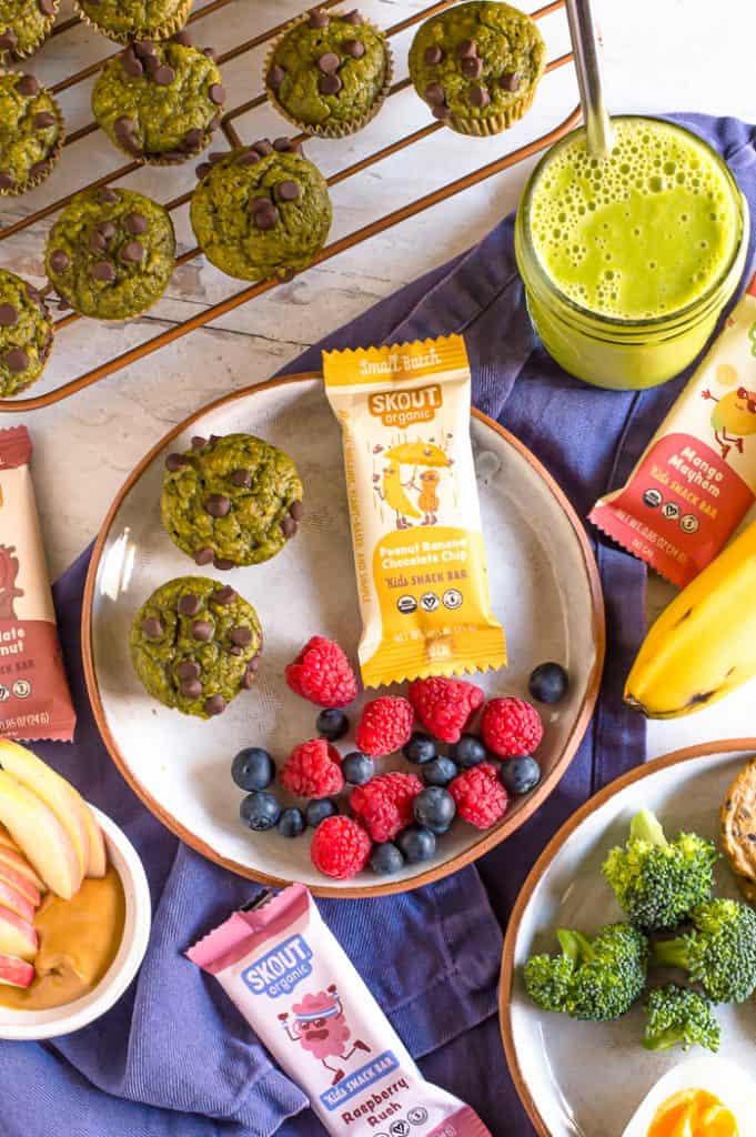 A spread of different healthy after school snack ideas for kids. There is a plate with two mini spinach muffins, blueberries and raspberries, and a Skout Organic bar. Next to it is a cooling rack of more muffins, a green smoothie, a plate of broccoli and more Skout organic bars.