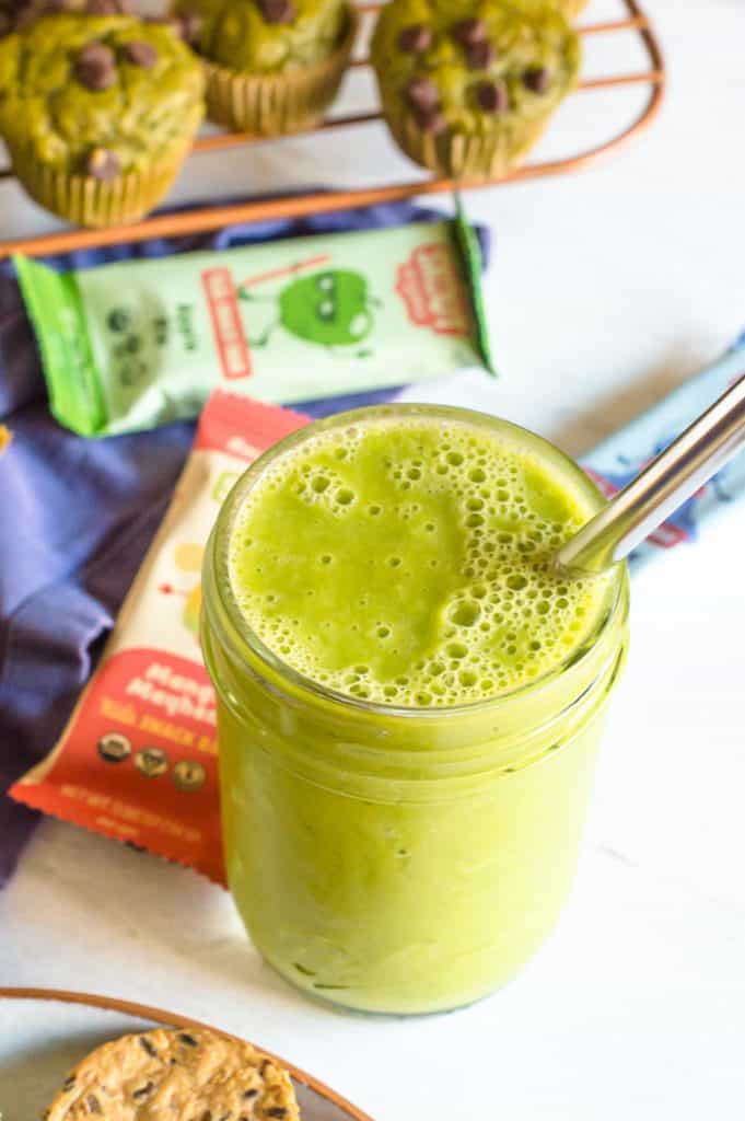 A green smoothie in a mason jar with a metal straw on a white table. There are Skout Organic bars scattered behind it on the table