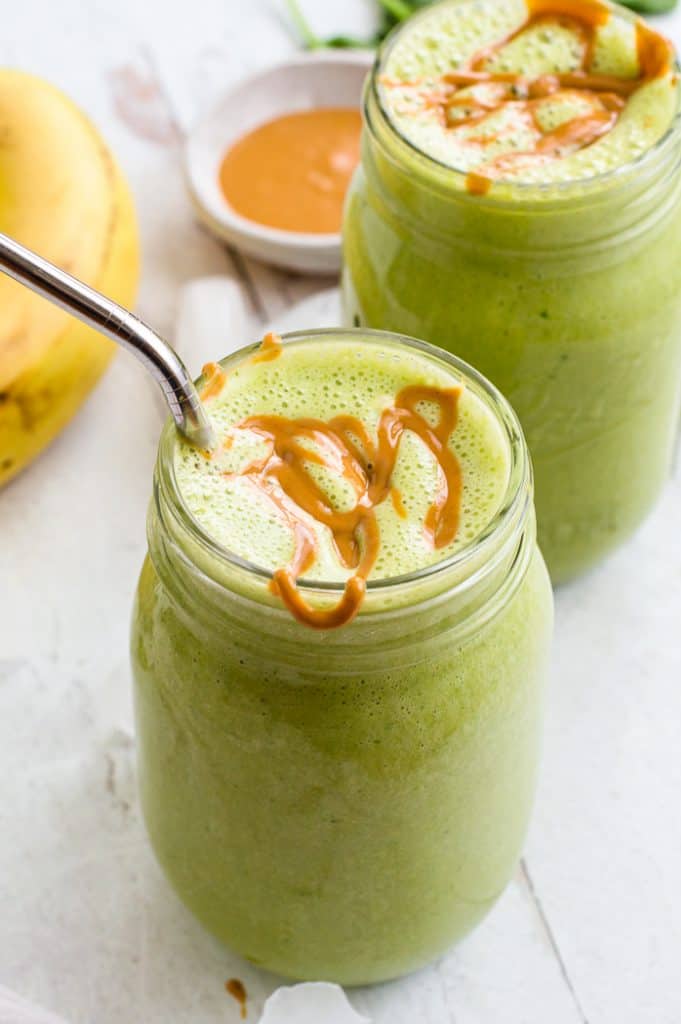 Spinach banana smoothie in two mason jars with peanut butter drizzled on top. One jar has a straw in it.