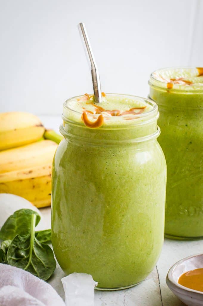 Banana spinach smoothie in two mason jars, topped with a drizzle of peanut butter. One jar has a metal straw in it. 