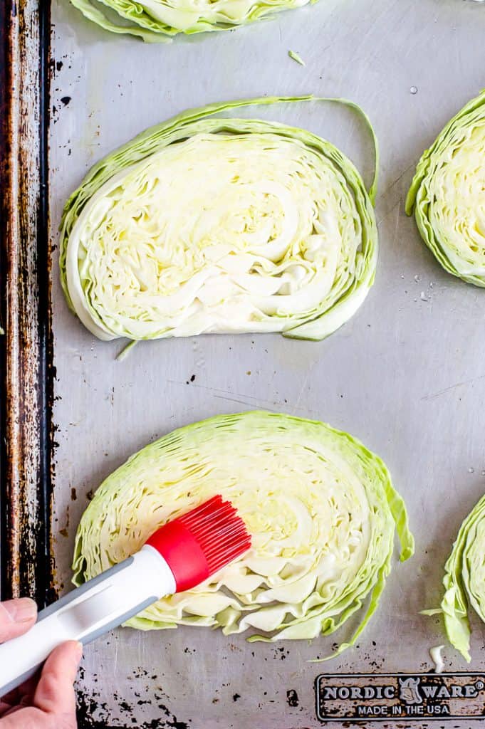 Cabbage steaks arranged on a baking sheet. A hand holding a pastry brush is brushing oil on one of the uncooked cabbaged steaks.