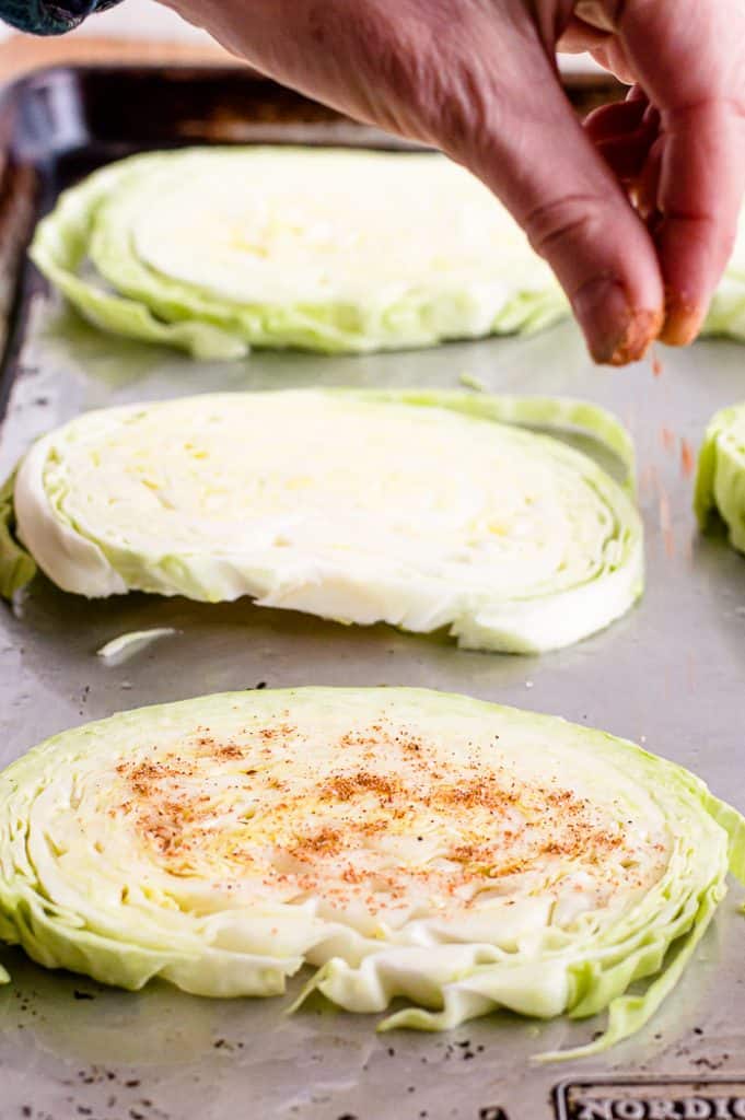 A hand sprinkling seasoning over uncooked cabbage steaks arranged on a metal baking sheet.
