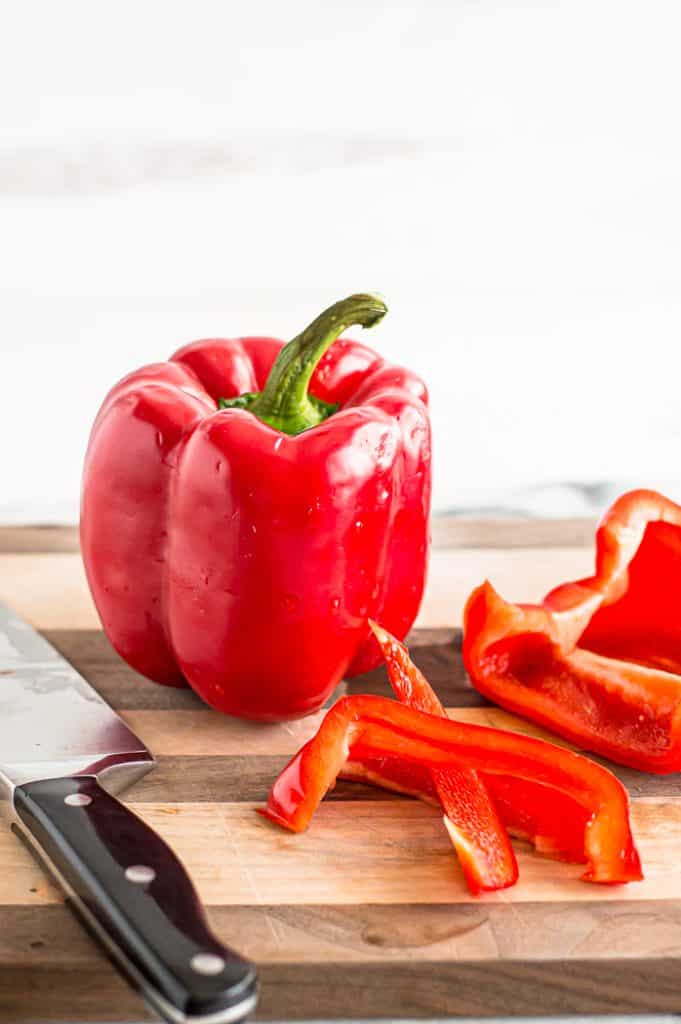 A red bell pepper on a cutting board with a knife. Another bell pepper that has been cut into slices sits next to it.