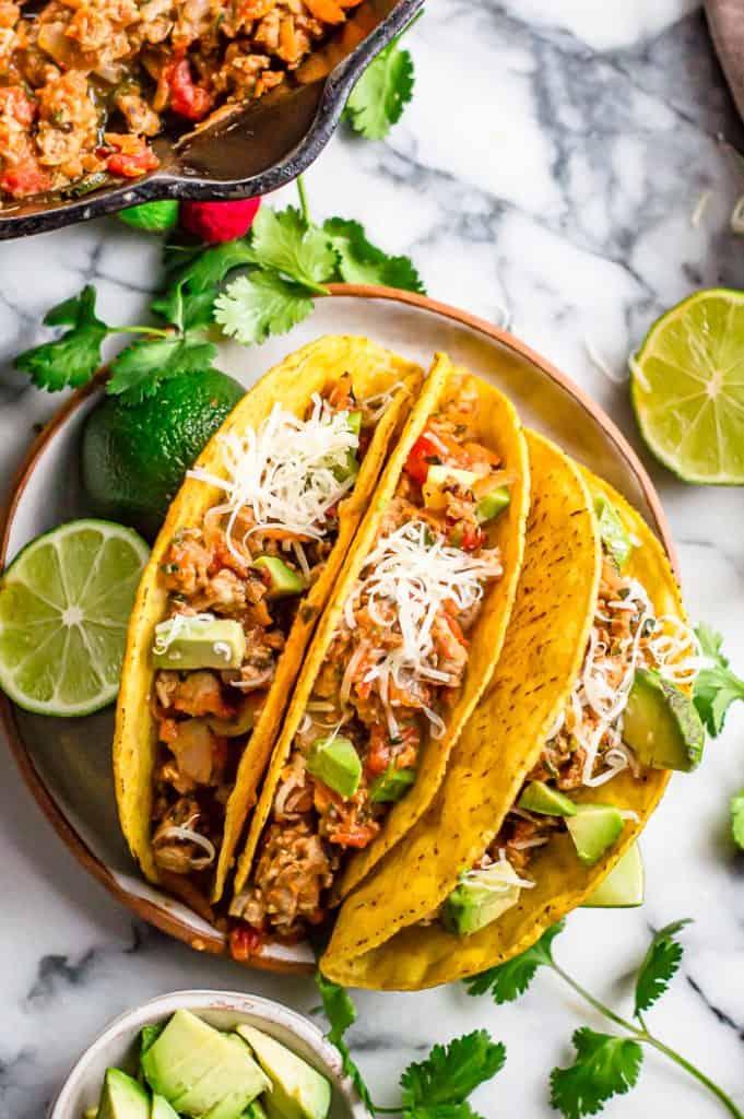 3 Veggie-loaded turkey tacos on a plate with cheese and diced avocado on top. There are limes on the plate with them and these is fresh cilantro and the skillet with the rest of the turkey meat on the table next to it.