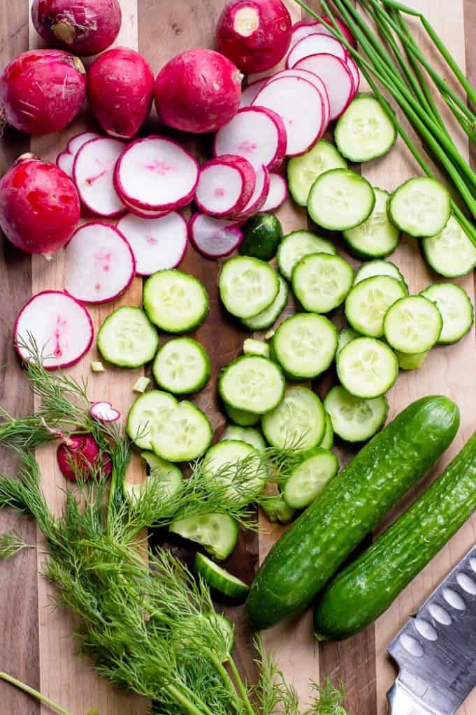 A wooden cutting board with sliced cucumbers, sliced radishes and fresh dill and chives with a chef's knife.