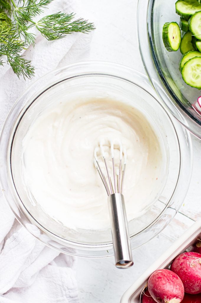 Creamy dill dressing in a bowl with a whisk on a white table. There is a bowl with cucumbers and another with radishes next to it. Fresh dill is in the corner.