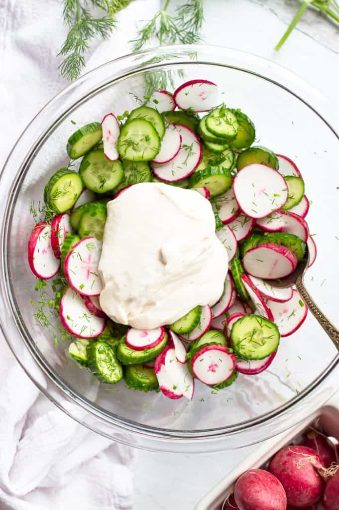 Cucumber radish salad in a bowl with a spoon with the creamy dill dressing poured on top but not mixed in yet.
